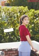 A woman in a red shirt and blue skirt leaning against a brick wall.